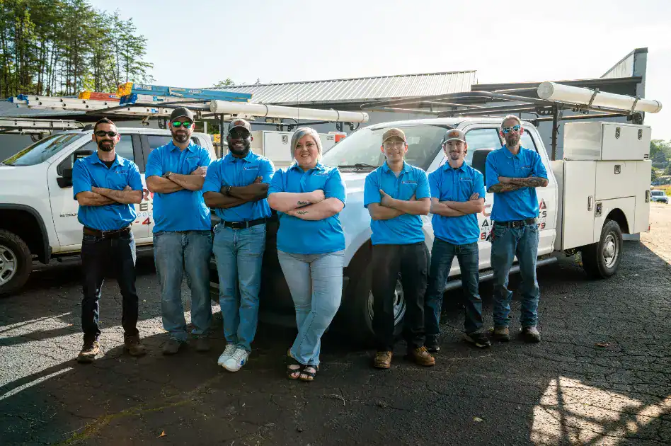 Seven people in blue shirts stand in front of white work trucks with ladders, posing for a group photo outdoors on a sunny day.